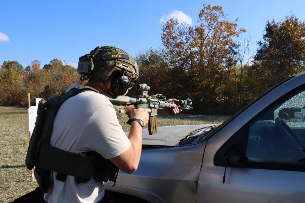 Competitor in tactical gear aiming a rifle beside a vehicle